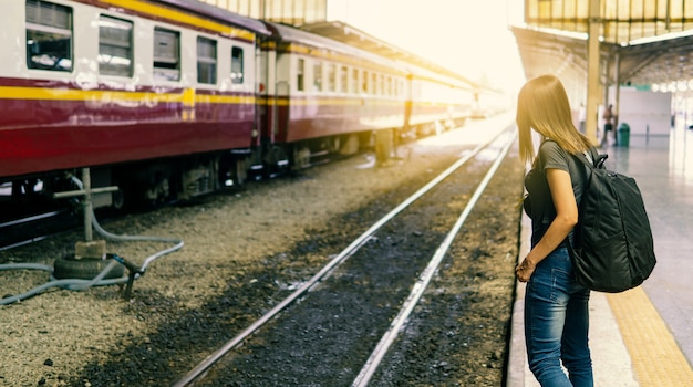 Rear view of young asian woman traveler with small backpack on the railway stantion.Waiting for train with transport on holiday trip.Travel concept