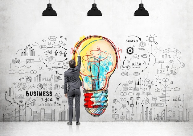 Rear view of a young businessman in a dark suit drawing a creative idea sketch on a concrete wall with a paintbrush.