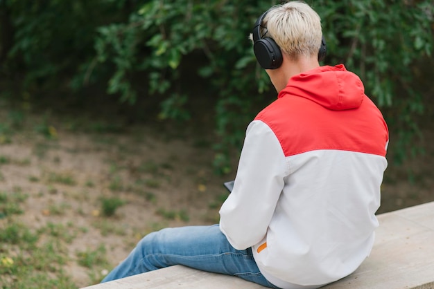 Rear view of young Caucasian blond guy sitting on wall and listening to music outdoors in park Lifestyle concept