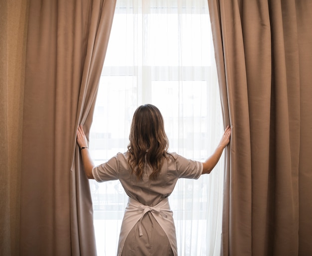 Rear view of a young maid opening curtains in hotel room