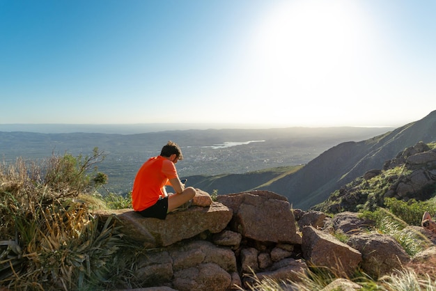 Rear View of Young Man Sitting on Rock in the Top of the Mountain Texting whit Smartphone