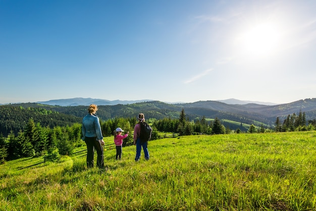 Rear view young mother and two daughters go down the hill overgrown with green grass. Gorgeous view of the forest growing on the mountains during trekking on a warm summer day