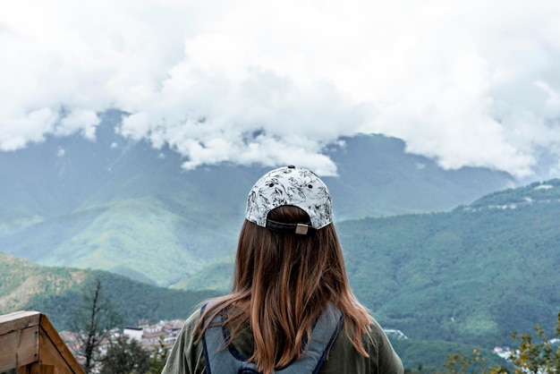 Rear view of young woman in cap with backpack plaid shirt looking at view of mountains cloudy sky