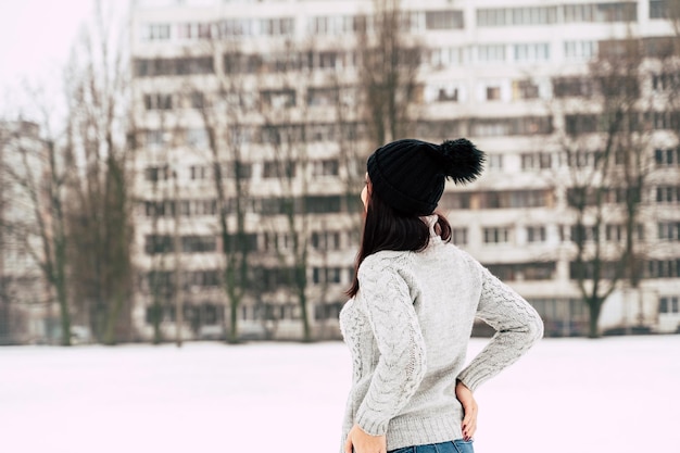 Rear view of young woman in gray knitted sweater and hat standing on street in winter season Female looking at highrise building in cloudy weather