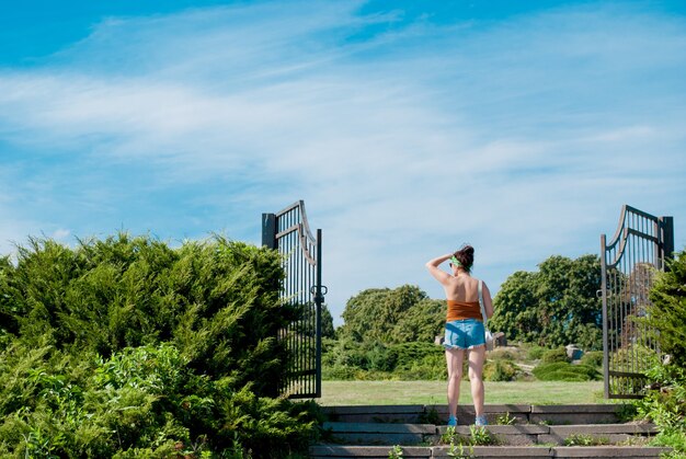 Rear view of a young woman looking into the distance at the entrance to the park in a large metal forged gate.