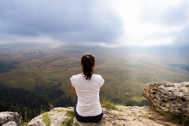 Rear view of a young woman sitting at the top of a mountain looking into the distance on the