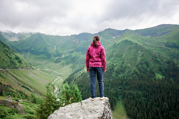 Rear view of young woman traveler standing on rock edge among magnificent green mountains