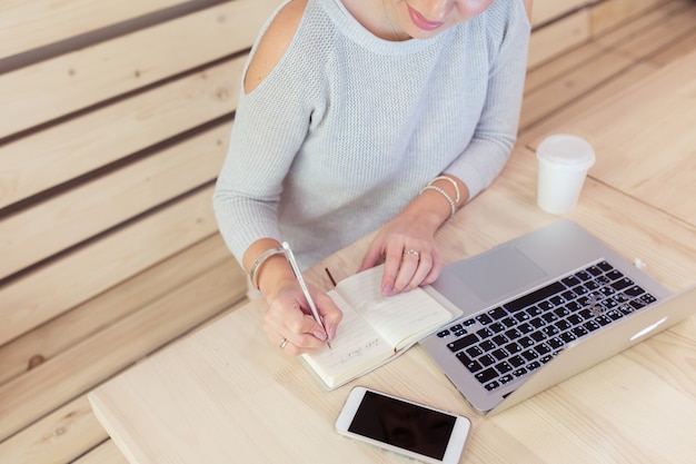 Rear view of young woman using laptop for surfing and browsing Internet, searching for required information, sitting at desk with mobile phone and notebook