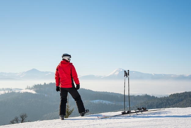 Rearview shot of a skier resting after the ride standing on top of the ski slope looking around enjoying breathtaking view of snowy mountains on sunny winter day