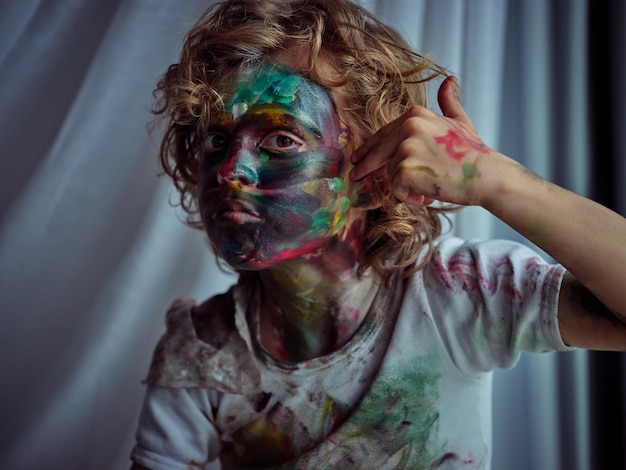 Rebellious dirty boy with curly hair smearing colorful paint on face while looking at camera with serious gaze in light studio