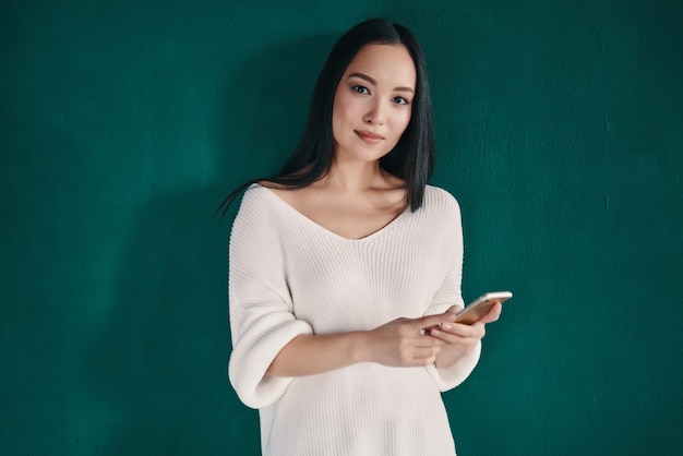 Receiving nice messages. Beautiful young woman using smart phone and smiling while standing against green wall