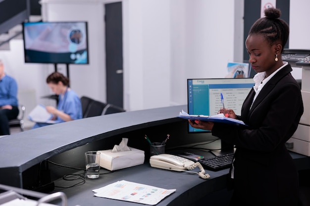 Receptionist in black suit holding clipboard checking medical documents while working at expertise in hospital waiting area. Reception worker is responsible for scheduling appointments