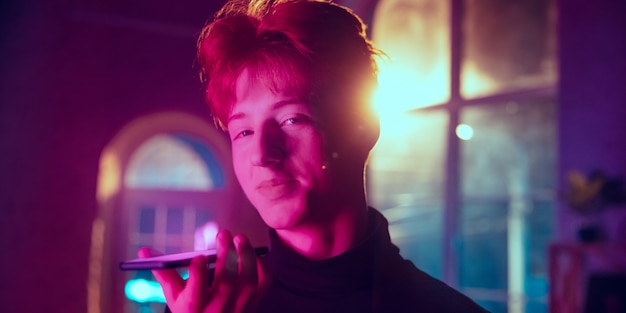 Recording voice message. Cinematic portrait of redhair man in neon lighted interior. Toned like cinema effects in purple-blue. Caucasian model using smartphone in colorful lights indoors. Flyer.