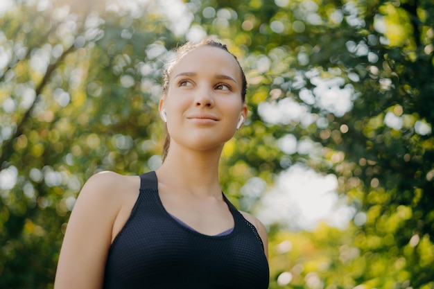 Recreation lifestyle and sport concept Thoughtful dark haired woman looks away with dreamy expression exercises outdoor in park during sunny summer day listens music in earphones has active life