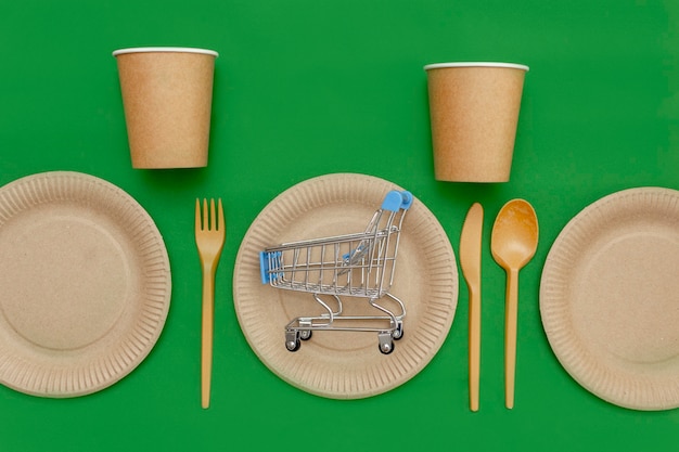 On a recyclable plate is a small shopping cart, next to a fork, spoon, knife, and a cup on a green background. Kitchen utensils are served on the table. Top view. Minimalist Style. Copy space.