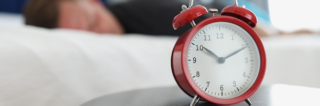 A red alarm clock stands on a round table in the bedroom against the background of a sleeping