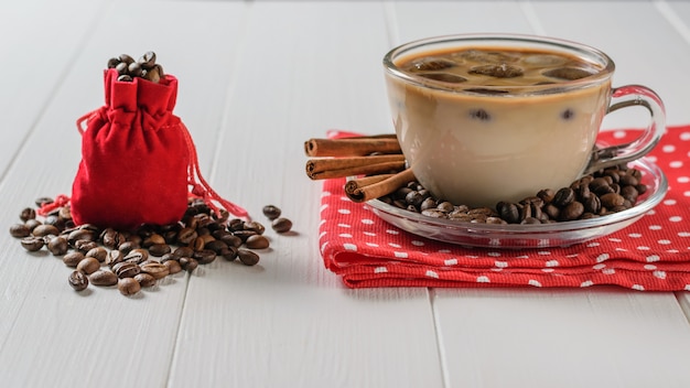 A red bag filled with coffee beans and a Cup of ice coffee and cinnamon on a white table.