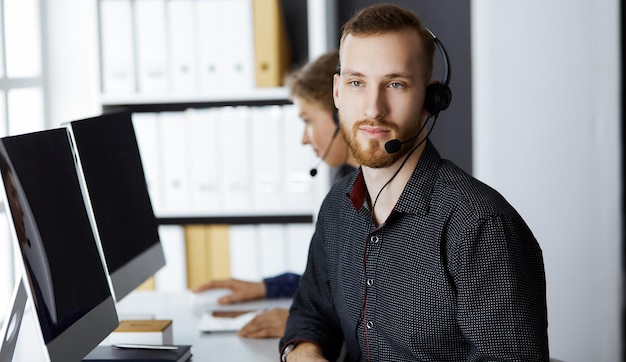 Red-bearded businessman talking by headset near his female colleague while sitting in modern office. Diverse people group in call center. Telemarketing and customer service.