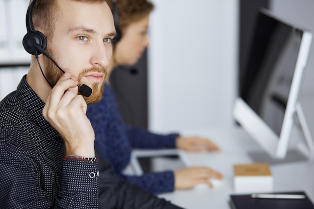 Red-bearded businessman talking by headset near his female colleague while sitting in modern office. Diverse people group in call center. Telemarketing and customer service.