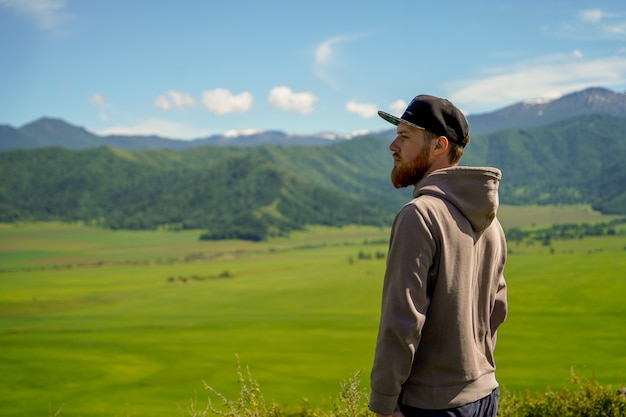 A red-bearded man in hoody standing in front of green field and mountains