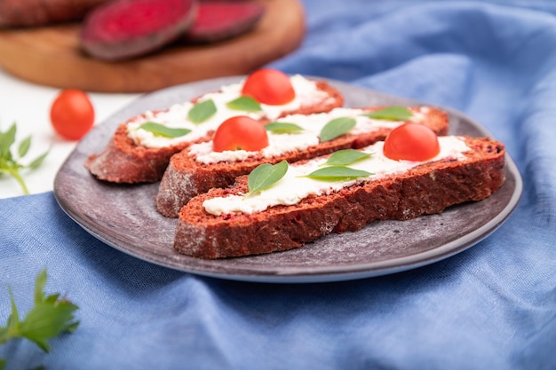 Red beet bread sandwiches with cream cheese and tomatoes on white concrete background and blue linen textile. Side view, selective focus, close up.