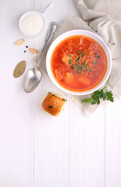 Red borscht soup in white bowl with sour cream and parsley, top view, on wooden white background.