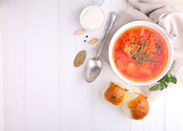 Red borscht soup in white bowl with sour cream and parsley, top view, on wooden white background