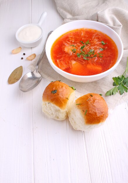 Red borscht soup in white bowl with sour cream and parsley, top view, on wooden white background