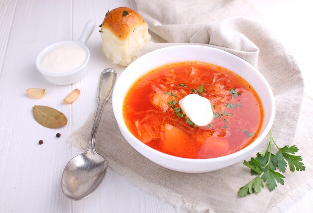 Red borscht soup in white bowl with sour cream and parsley, top view, on wooden white background