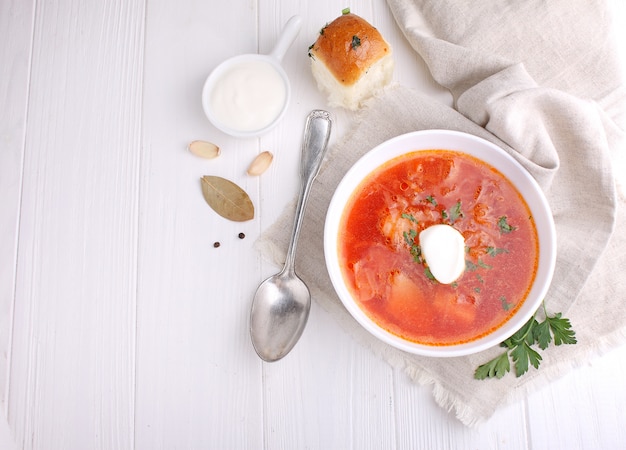 Red borscht soup in white bowl with sour cream and parsley, top view, on wooden white background