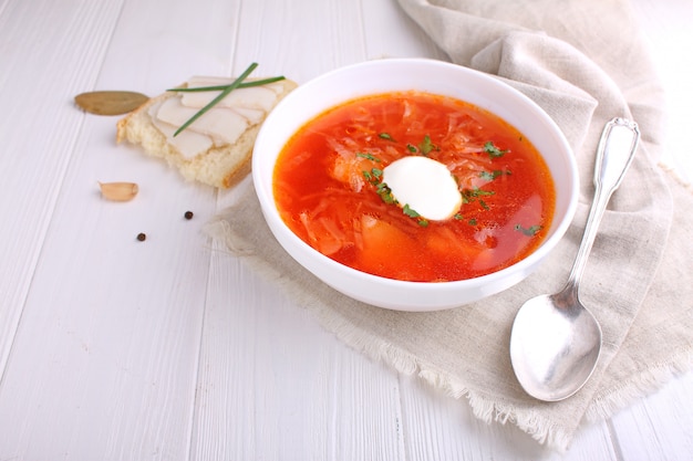 Red borscht soup in white bowl with sour cream and parsley, top view, on wooden white background.