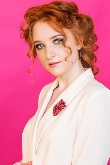 A red brooch in the shape of an anatomical heart on a redhaired girl's white jacket The girl poses on a bright pink background