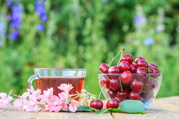 Photo red cherry fruits with peduncles in glass bowl and cup of tea on old wooden boards.