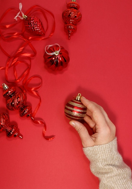 Red Christmas balls on a red background top view. A female hand holds a festive New Year's decor, flat lay.