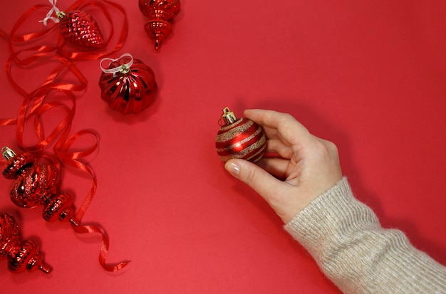 Red Christmas balls on a red background top view. A female hand holds a festive New Year's decor, flat lay.