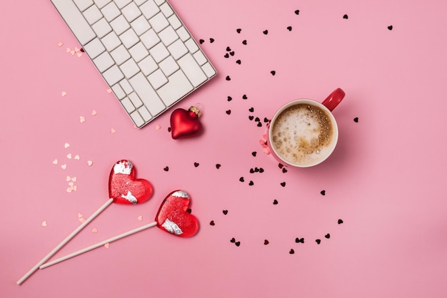 Photo red cup of coffee white keyboard sparkling red confetti two lollipop in shape of heart pink background blogger workplace on valentine's day holiday concept