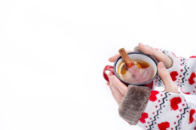 A red cup of hot tea with cinnamon in hands with warm mitts on a background of white snow