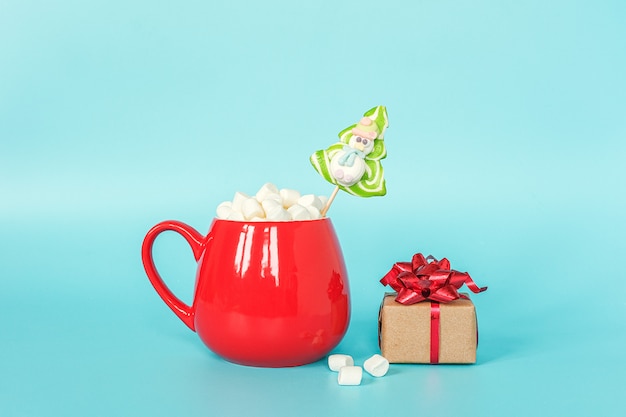 Red cup of marshmallow with green lollipop christmas tree and gift box on blue background.