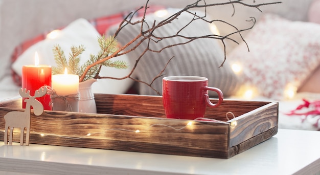 Red cup of tea on tray with burning candles sofa with pillows. Cozy home concept