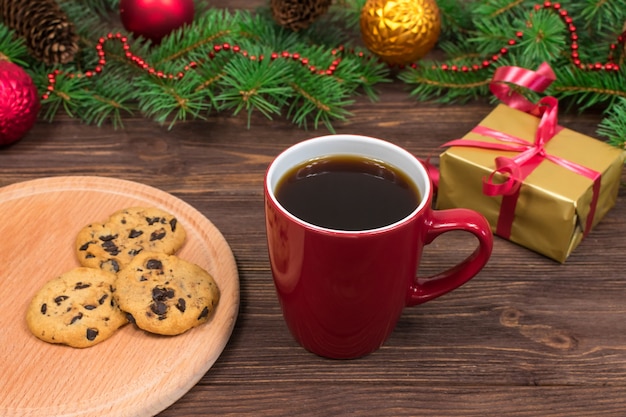 Red cup with tea, coffee with cookies and marshmallows on a wooden table against the background of a New Year tree with Christmas decorations.