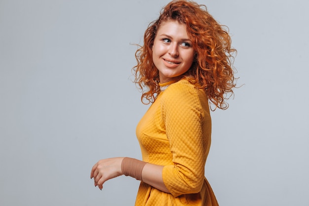 Red curly girl in a yellow dress on a light gray background smiling