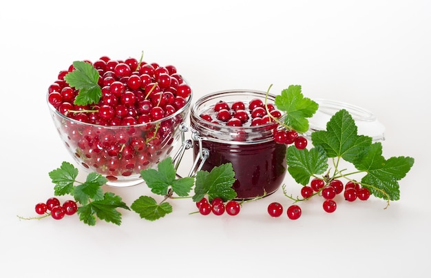 Red currant jam in a jar and ripe berries in a glass cup with green leaves on a white background.