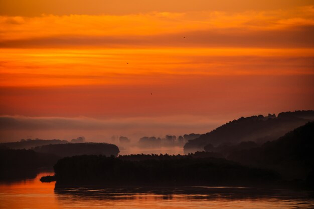 Red dawn sky reflected on water in valley of river. Morning haze in distance above forest on shore. Birds flying in sky at sunrise. Fog on riverbank.