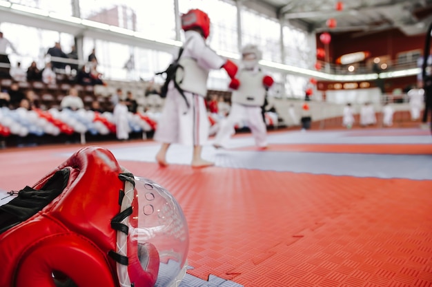 Red fighting helmet with clear plastic mask on floor Two boys in kimono in karate fight battle blur