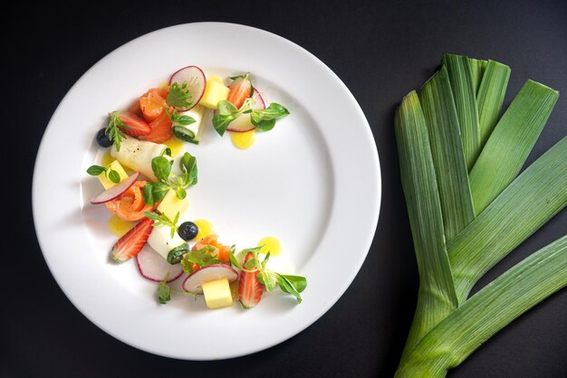 Red fish vegetables and fruit salad on a white plate Fish fillet rolls radish zucchini rolls microgreens strawberry wedges mango cubes juice drops leek Top view on a dark black background