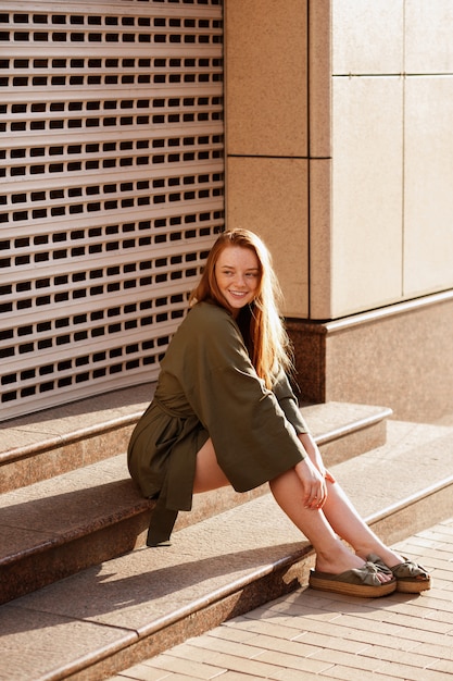 Red-haired girl with freckles in green clothes sits on the steps on a walk. summer mood