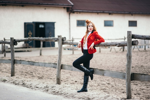 Red-haired jockey woman in a red cardigan and black high boots stares at the fence