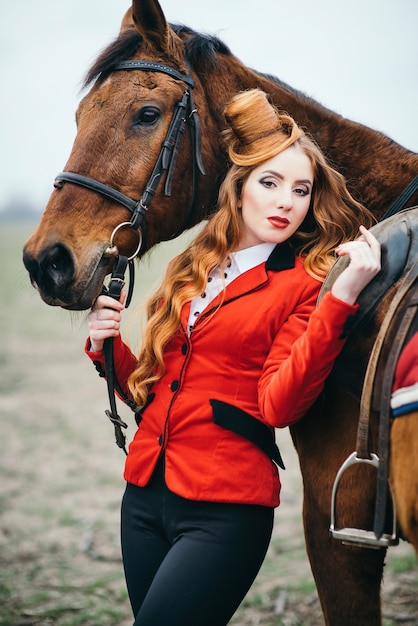 Red-haired jockey woman in a red cardigan and black high boots with a horse for a walk