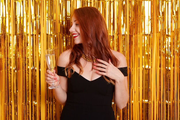 Red haired smiling woman with glass of wine looking aside, wearing black dress with bare shoulders, stands against wall with golden tinsel