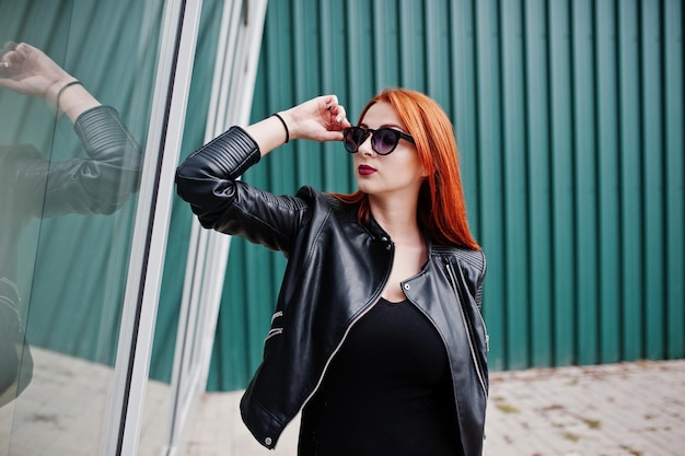 Red haired stylish girl in sunglasses wear in black against large windows Street fashion portrait